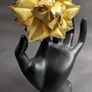 May include: A gold-colored flower brooch with layered petals. The brooch is displayed on a black hand-shaped sculpture against a gray background. The flower's petals have a textured appearance, reflecting light.