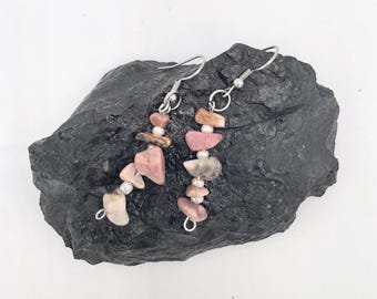 May include: Dangle earrings with pink, beige, and grey stones, strung on silver-coloured hooks with small beads. The earrings are displayed on a dark grey, textured surface. The stones are of varying shapes and sizes.
