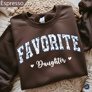 May include: A brown sweatshirt with the word "FAVORITE" in white block letters filled with a blue floral pattern. Below, the word "Daughter" is written in white script, with a white heart on each side.