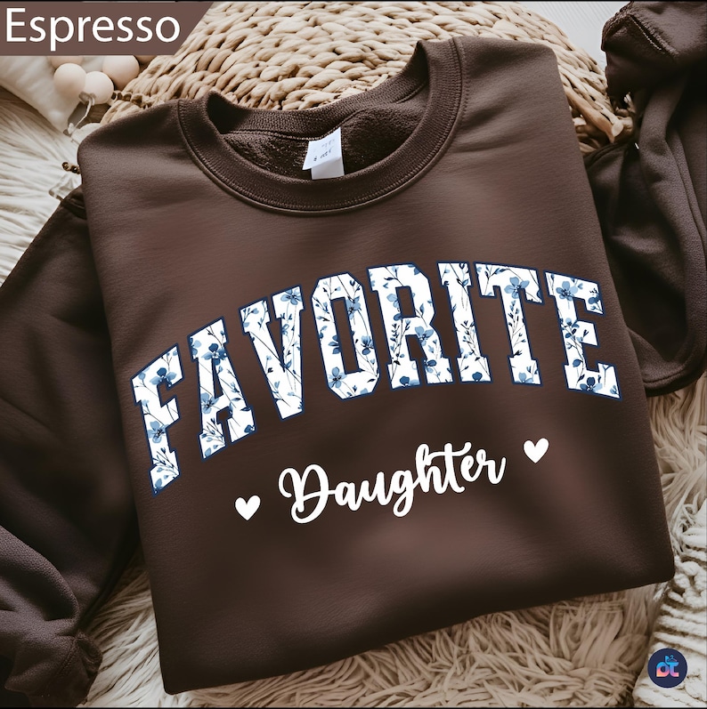 May include: A brown sweatshirt with the word "FAVORITE" in white block letters filled with a blue floral pattern. Below, the word "Daughter" is written in white script, with a white heart on each side.