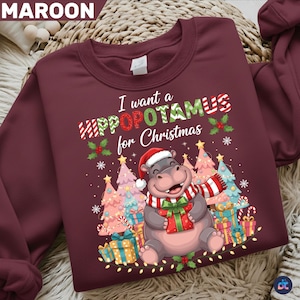May include: Maroon Christmas sweatshirt with the text "I want a Hippopotamus for Christmas." The design features a cartoon hippo wearing a Santa hat and scarf, holding a gift, surrounded by Christmas trees and presents.