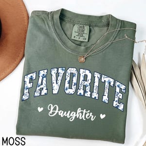 May include: Moss green Comfort Colors t-shirt with the words "FAVORITE Daughter" in white script and floral print. A gold necklace with a pendant rests on the shirt. The shirt is folded and displayed with a brown hat.