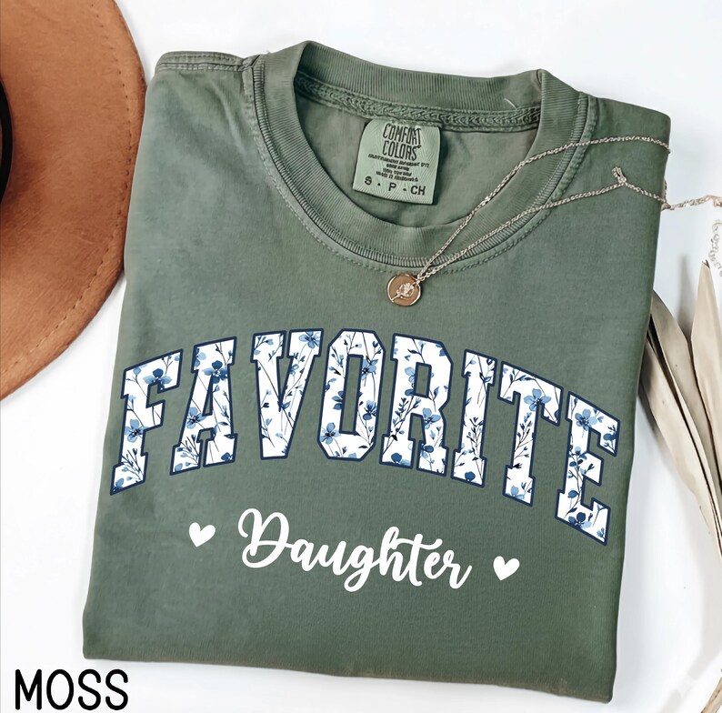 May include: Moss green Comfort Colors t-shirt with the words "FAVORITE Daughter" in white script and floral print. A gold necklace with a pendant rests on the shirt. The shirt is folded and displayed with a brown hat.