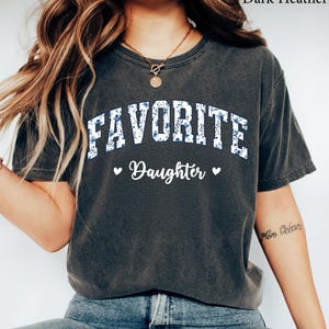 May include: Dark heather gray t-shirt with the word "FAVORITE" in white block letters filled with a blue floral pattern. Below, the word "Daughter" is written in script, with a heart on each side.