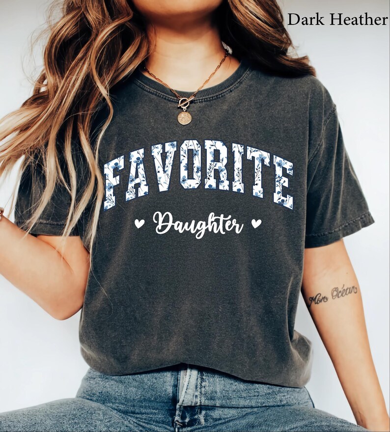 May include: Dark heather gray t-shirt with the word "FAVORITE" in white block letters filled with a blue floral pattern. Below, the word "Daughter" is written in script, with a heart on each side.