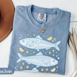 May include: A blue jean-coloured t-shirt featuring a fish graphic. The design includes two detailed fish with floral patterns, stars, and lemon slices. A gold necklace with a pendant rests on the shirt. The shirt has a Comfort Colors tag.