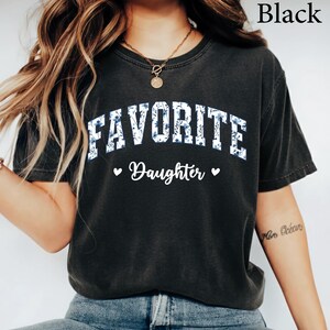 May include: Black t-shirt with the text "FAVORITE Daughter" in white script and blue floral print. The word "FAVORITE" is in a collegiate style font. Two small hearts flank the word "Daughter".