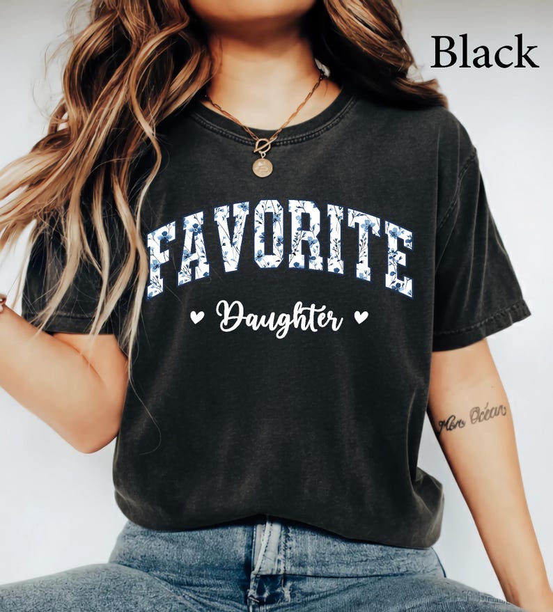May include: Black t-shirt with the text "FAVORITE Daughter" in white script and blue floral print. The word "FAVORITE" is in a collegiate style font. Two small hearts flank the word "Daughter".