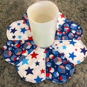 Può includere: Una candela a colonna bianca è posizionata su un sottobicchiere patriottico a forma di fiore. Il sottobicchiere presenta un design patchwork con tessuti bianchi, blu navy e rossi. Il tessuto è decorato con stelle e forme geometriche.