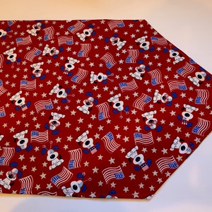 Può includere: Tovaglia da tavolo rossa con un motivo di cani bianchi che indossano cappelli rosso, bianco e blu e tengono bandiere americane. Lo sfondo è pieno di stelle bianche.