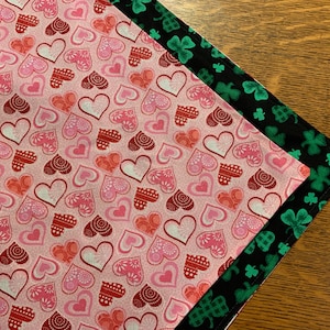 Könnte beinhalten: Zwei Stoffquadrate, eines mit rosa Hintergrund und einem Muster aus roten und rosa Herzen, das andere mit schwarzem Hintergrund und einem Muster aus grünen Kleeblättern.