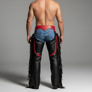 K&ouml;nnte beinhalten: Schwarze Lederchaps mit roten Akzenten und Fransen. Die Chaps werden &uuml;ber einer blauen Jeans getragen und haben einen roten G&uuml;rtel. Das Design umfasst dekorative N&auml;hte und einen klassischen Western-Stil.
