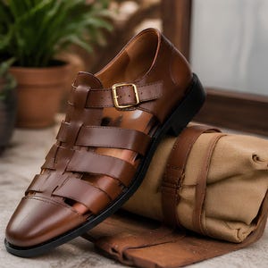 May include: A brown leather fisherman sandal with a closed toe, a series of wide straps across the upper, and a gold-toned metal buckle closure on the side. The shoe features a solid black sole and is positioned next to a folded tan fabric roll.