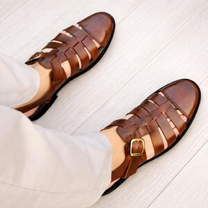 May include: A pair of brown leather fisherman sandals featuring a closed-toe design with woven straps, a secure ankle strap with a gold-tone metal buckle, and a black sole. The sandals are worn with light-colored trousers, showcasing the detailed construction.