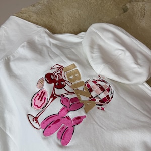 Puede incluir: Sudadera con capucha blanca con un diseño bordado colorido. El diseño incluye un cóctel, un perro globo rosa y la palabra "BIZZA" en letras doradas. Un globo rosa y blanco a cuadros también forma parte del diseño.