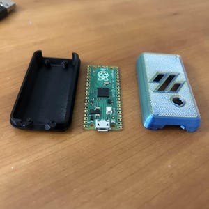 Puede incluir: Una carcasa negra impresa en 3D abierta, una placa de circuito verde con un logotipo de Raspberry Pi y una carcasa azul y gris impresa en 3D con un diseño geométrico se muestran sobre una superficie de madera. Un cable USB es visible en el fondo.