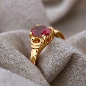 May include: A gold ring with a large, oval-shaped, red gemstone. The ring's band and setting are a bright gold color. The ring is set against a neutral-toned fabric, highlighting the gemstone's color and the ring's design.