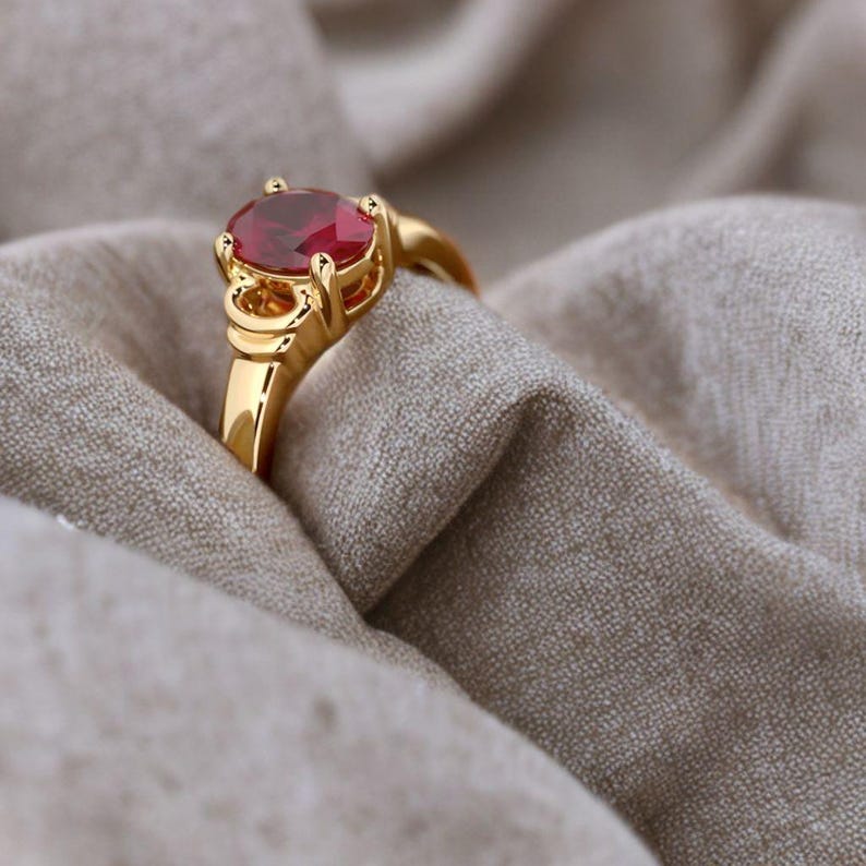 May include: A gold ring with a large, oval-shaped, red gemstone. The ring's band and setting are a bright gold color. The ring is set against a neutral-toned fabric, highlighting the gemstone's color and the ring's design.
