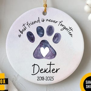 May include: A round ceramic ornament with a paw print design. The paw print is purple with a white heart in the center. The text reads "a best friend is never forgotten" and the name "Dexter" with the years 2018-2025.