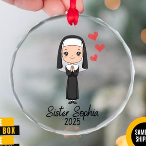May include: A clear, round ornament with a cartoon nun design. The nun is wearing a black habit and white wimple, with three red hearts above her head. The text "Sister Sophia 2025" is printed below the nun.