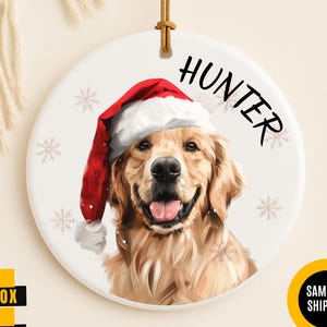 May include: A round white ceramic ornament featuring a golden retriever wearing a red and white Santa hat. The name HUNTER is printed in black text above the dog. The ornament includes a brown leather hanging cord and light snowflake accents.