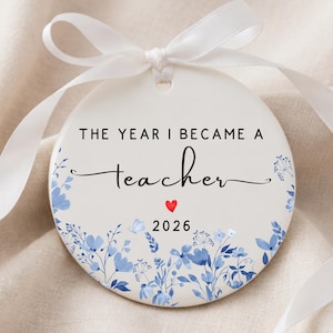 May include: A round white ceramic ornament featuring a white ribbon tied at the top. The text reads THE YEAR I BECAME A teacher, with a small red heart and the year 2026 below. The bottom edge is decorated with a blue floral botanical illustration.