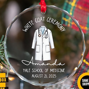 May include: A clear glass ornament with a red ribbon, featuring a white coat illustration and the text "WHITE COAT CEREMONY." It includes the name "Amanda," and details "YALE SCHOOL OF MEDICINE AUGUST 21, 2025."