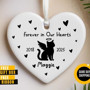 May include: White heart-shaped ornament with a black cat silhouette, angel wings, and halo. Text reads "Forever in Our Hearts," with the years 2018 and 2025. Small black hearts surround the text and cat. A white ribbon is attached.
