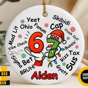 May include: A round white ceramic ornament featuring the number 67 in red and green, with a Santa hat on the 7 labeled 2026. The design includes colorful string lights, red and green snowflakes, and slang terms like Yeet, Ohio, Fanum, Skibidi, Cap, and the name Aiden.