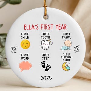 May include: A white ceramic disc ornament with a gold hanging loop. It displays the text "Ella's First Year" and baby milestone illustrations: smile, tooth, crawl, first word, step, and sleeping through the night. The year 2025 is printed at the bottom.