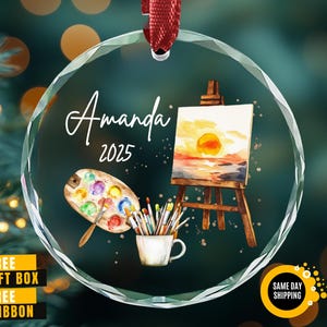 May include: A clear, round Christmas ornament with a watercolor painting theme. The ornament features a painting of a sunset on an easel, a palette of paints, and a cup of paintbrushes. The name "Amanda" and the year "2025" are written on the ornament.