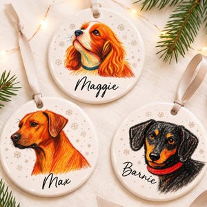 May include: Three round white ceramic ornaments with satin ribbons. Each features a colored pencil-style dog portrait and a name: Maggie, a golden spaniel; Max, a reddish-brown dog; and Barnie, a black and tan dachshund with a red collar.