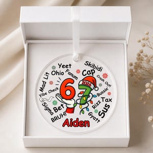May include: A round, clear acrylic ornament featuring the number 67 in red and green, with a Santa hat on the 7 and the year 2026. The design includes various slang terms like Yeet, Ohio, and Skibidi, with the name Aiden in red at the bottom.