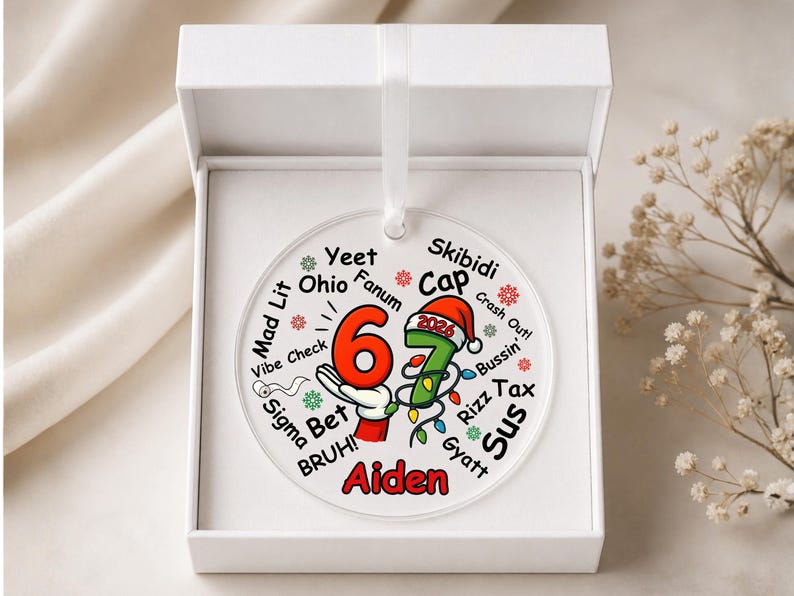 May include: A round, clear acrylic ornament featuring the number 67 in red and green, with a Santa hat on the 7 and the year 2026. The design includes various slang terms like Yeet, Ohio, and Skibidi, with the name Aiden in red at the bottom.