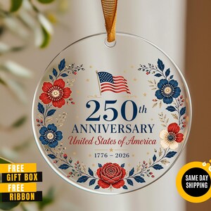May include: A clear, round ornament with a gold ribbon for hanging. The ornament features the text "250th ANNIVERSARY United States of America 1776-2026" with a US flag and floral design in red, white, and blue. The words "FREE GIFT BOX FREE RIBBON" are also visible.