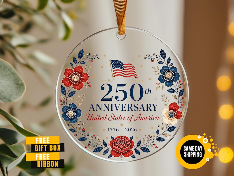 May include: A clear, round ornament with a gold ribbon for hanging. The ornament features the text "250th ANNIVERSARY United States of America 1776-2026" with a US flag and floral design in red, white, and blue. The words "FREE GIFT BOX FREE RIBBON" are also visible.