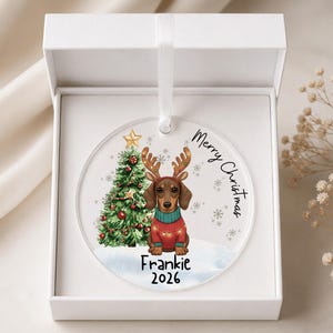 May include: A round clear acrylic Christmas ornament featuring a dachshund wearing reindeer antlers and a red sweater, a decorated Christmas tree, and the text Merry Christmas, Frankie, and 2026, presented inside a white gift box with a ribbon.