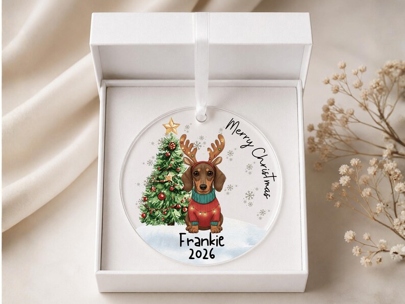 May include: A round clear acrylic Christmas ornament featuring a dachshund wearing reindeer antlers and a red sweater, a decorated Christmas tree, and the text Merry Christmas, Frankie, and 2026, presented inside a white gift box with a ribbon.