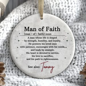 May include: A round, off-white ornament with the text "Man of Faith" and a definition. The ornament has a beige ribbon for hanging and is set against a white, textured background. The name "Tommy" is written in cursive.