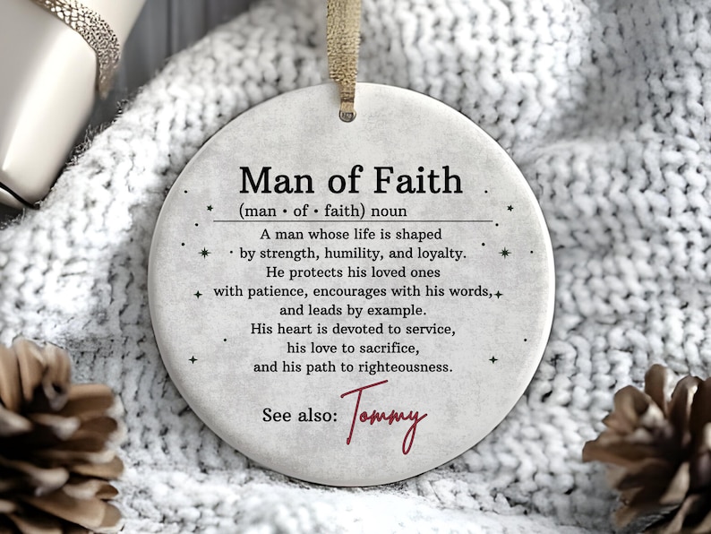 May include: A round, off-white ornament with the text "Man of Faith" and a definition. The ornament has a beige ribbon for hanging and is set against a white, textured background. The name "Tommy" is written in cursive.