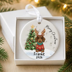 May include: A round white ceramic Christmas ornament featuring a brown dachshund wearing reindeer antlers and a red sweater. Next to the dog is a decorated Christmas tree. The text reads Merry Christmas, Frankie, and 2026. A white ribbon is attached.