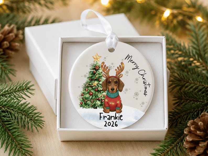 May include: A round white ceramic Christmas ornament featuring a brown dachshund wearing reindeer antlers and a red sweater. Next to the dog is a decorated Christmas tree. The text reads Merry Christmas, Frankie, and 2026. A white ribbon is attached.