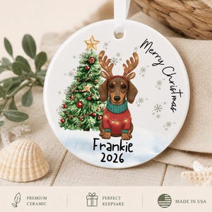 May include: A round white ceramic ornament featuring a brown dachshund wearing reindeer antlers and a red sweater with string lights. Next to the dog is a decorated Christmas tree with a gold star. The text reads Merry Christmas, Frankie, and 2026.