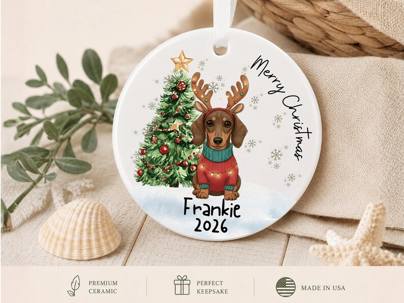 May include: A round white ceramic ornament featuring a brown dachshund wearing reindeer antlers and a red sweater with string lights. Next to the dog is a decorated Christmas tree with a gold star. The text reads Merry Christmas, Frankie, and 2026.