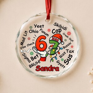 May include: A round, clear glass Christmas ornament with a red ribbon hanger. The design features the number 67, a cartoon hand, a Santa hat with 2026, and the name Sandra. It is surrounded by slang words like Yeet, Ohio, Fanum, Skibidi, Cap, and Rizz.