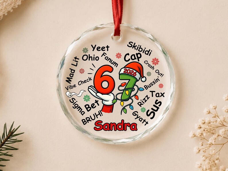 May include: A round, clear glass Christmas ornament with a red ribbon hanger. The design features the number 67, a cartoon hand, a Santa hat with 2026, and the name Sandra. It is surrounded by slang words like Yeet, Ohio, Fanum, Skibidi, Cap, and Rizz.