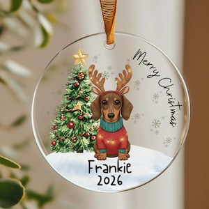 May include: A round clear acrylic Christmas ornament featuring a brown dachshund wearing a red sweater and reindeer antlers, a decorated green Christmas tree, and the text Merry Christmas, Frankie, and 2026, suspended by a gold ribbon.