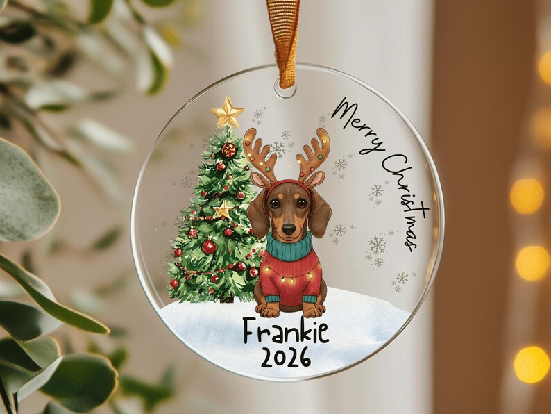 May include: A round clear acrylic Christmas ornament featuring a brown dachshund wearing a red sweater and reindeer antlers, a decorated green Christmas tree, and the text Merry Christmas, Frankie, and 2026, suspended by a gold ribbon.