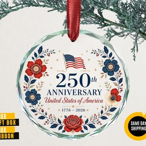 May include: A clear, round glass ornament with a red ribbon. The ornament features a floral wreath design in red and blue, surrounding the text "250th Anniversary United States of America 1776-2026" and an American flag.