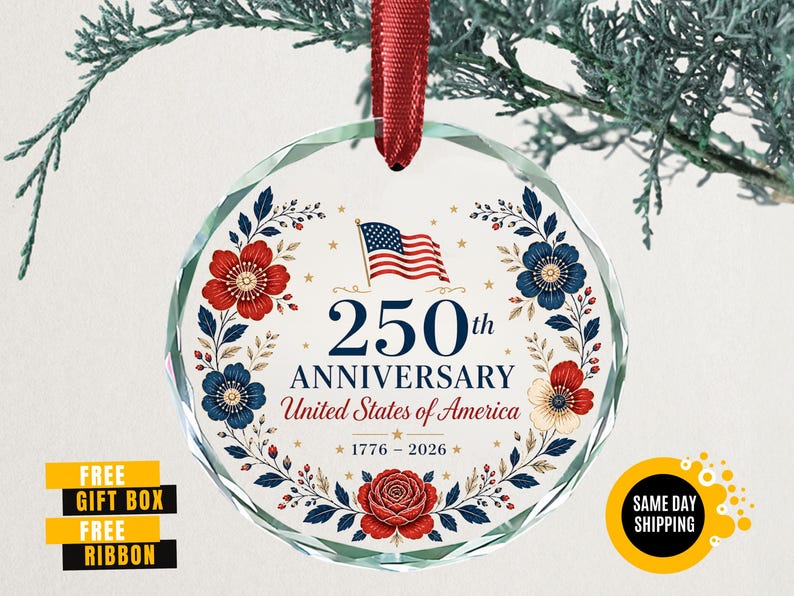 May include: A clear, round glass ornament with a red ribbon. The ornament features a floral wreath design in red and blue, surrounding the text "250th Anniversary United States of America 1776-2026" and an American flag.